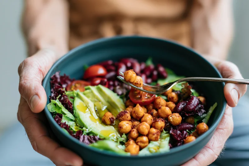 Hands holding a dark ceramic bowl filled with fresh salad of chickpeas, avocado, tomatoes, and lettuce