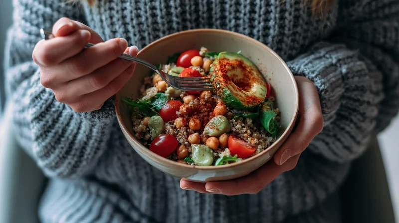 Woman holding a ceramic bowl of quinoa salad with avocado and cherry tomatoes, eating with a fork, indoors in a grey chunky knit sweater