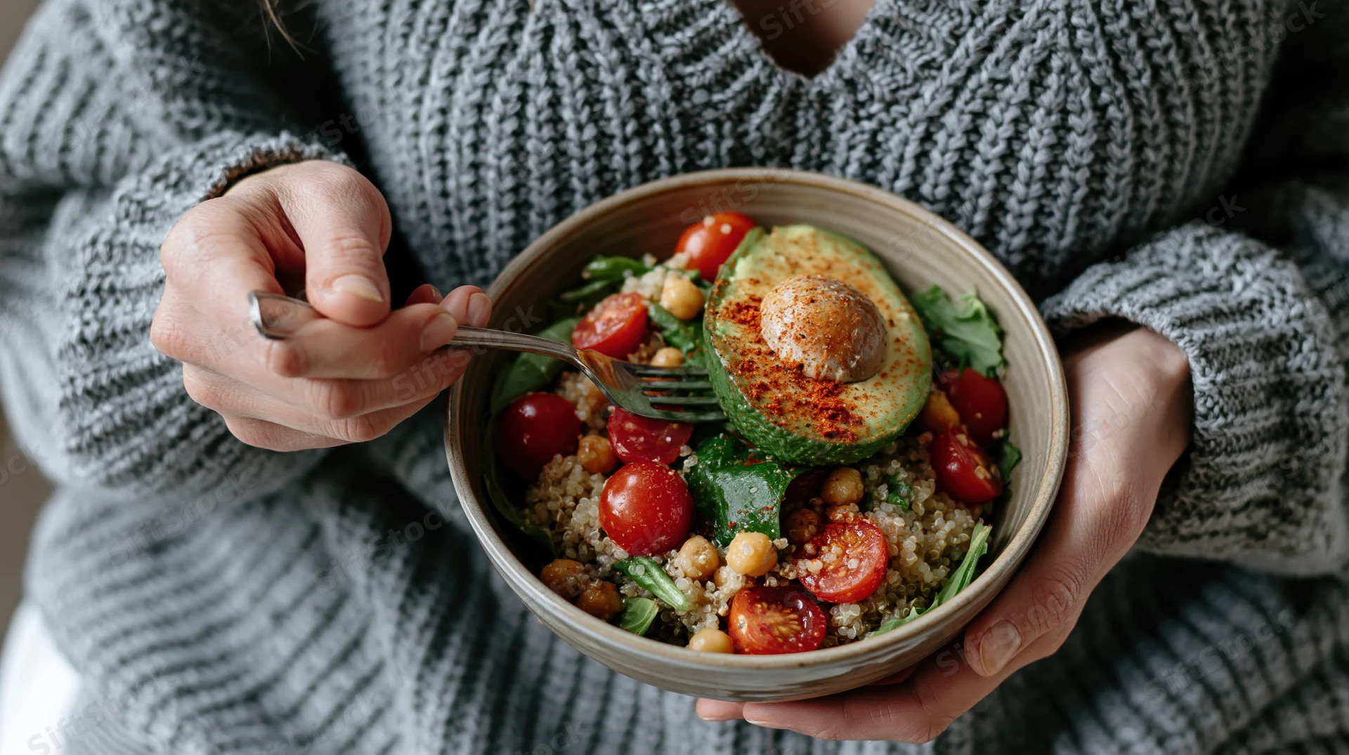 Woman holding a ceramic bowl of quinoa salad with avocado and cherry tomatoes, eating with a fork