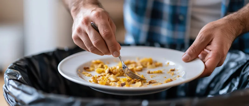 Man scraping leftover food from a white plate into a trash bin