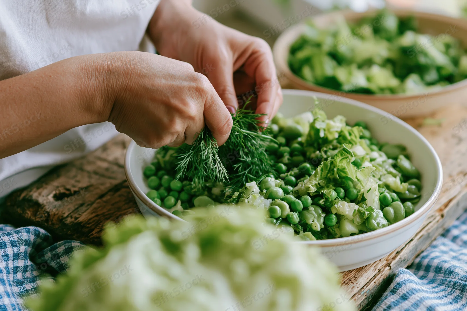 Hands adding fresh dill to a white bowl of green salad on a wooden table