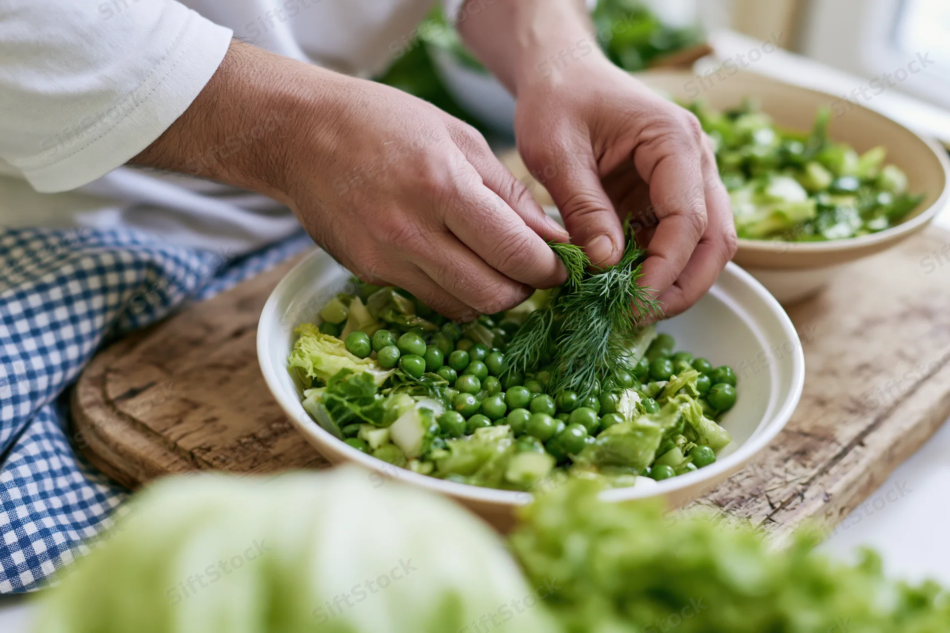 Hands adding fresh dill to a white bowl of green salad on a wooden cutting board