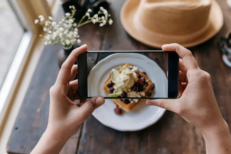 Hands holding smartphone horizontally photographing waffle dessert on wooden table near window