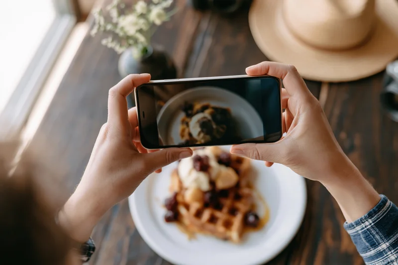Hands holding smartphone horizontally photographing waffle dessert on wooden table