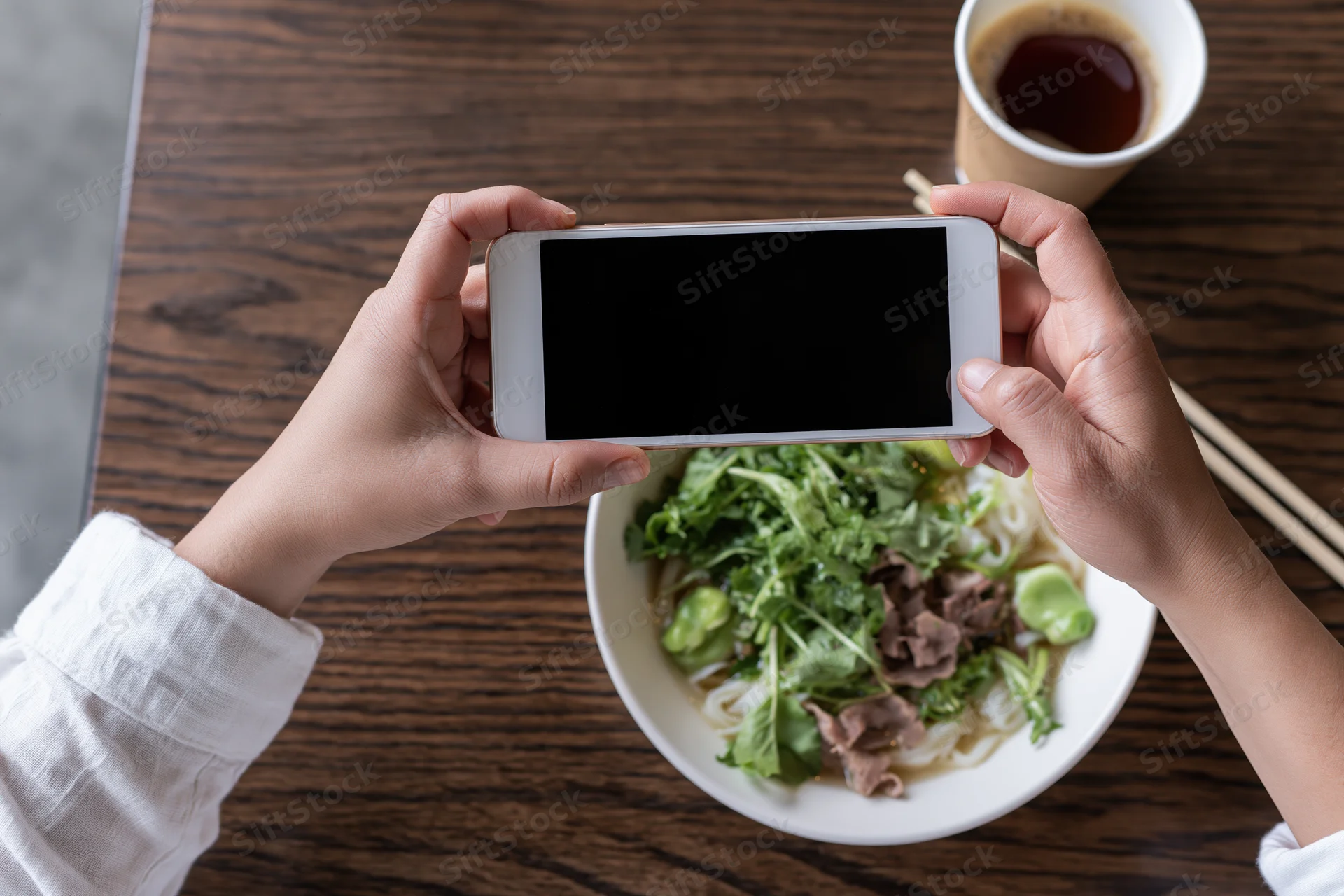 Hands holding smartphone overhead photographing bowl of noodles with greens and beef on wooden table