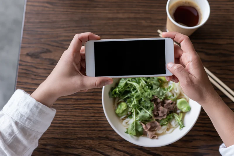 Hands holding smartphone overhead photographing bowl of noodles with greens and beef on wooden table