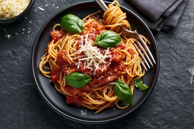 Spaghetti with tomato sauce and grated parmesan on a black plate, garnished with basil leaves, served with fork on dark stone surface