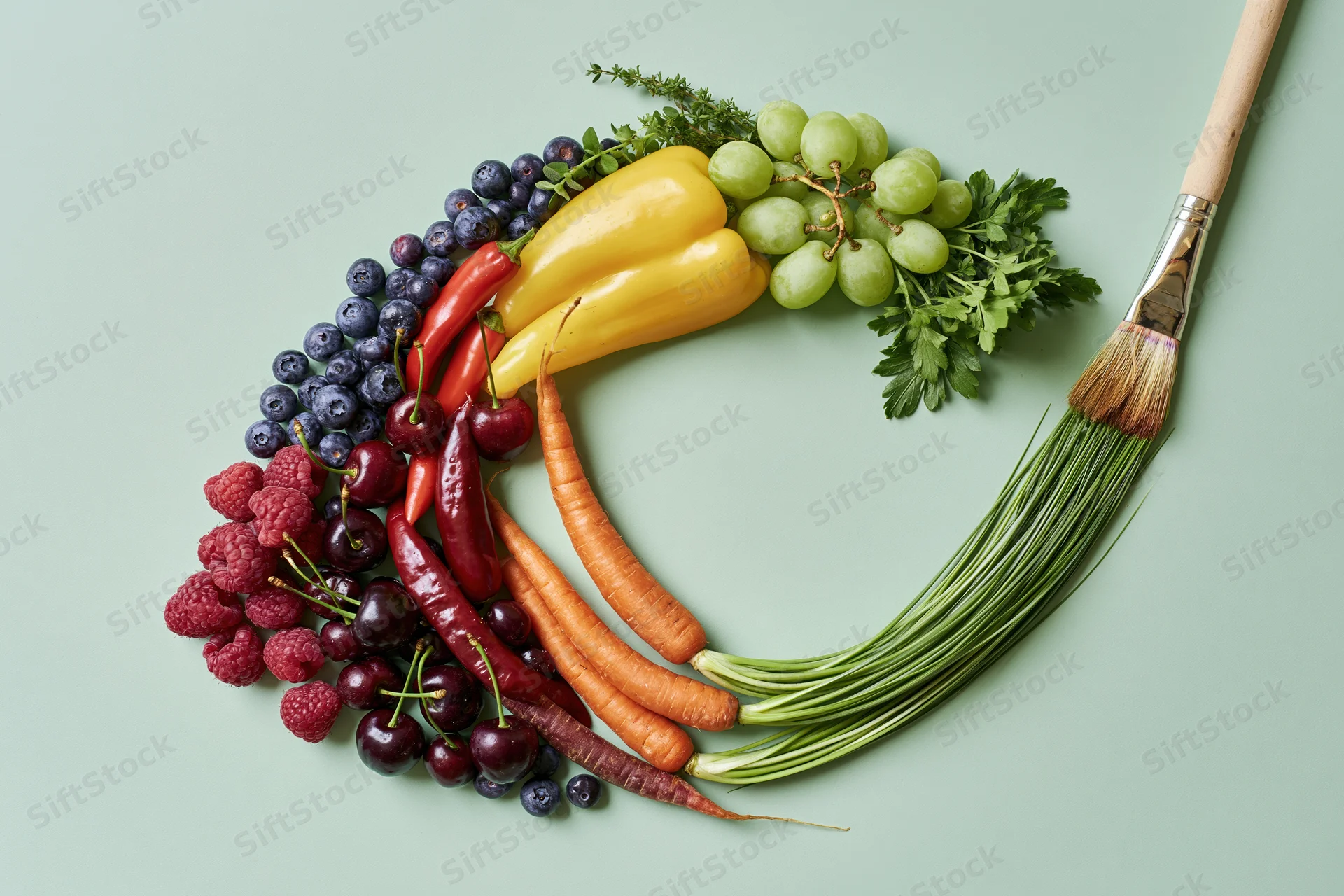 Rainbow arc of fresh fruits and vegetables arranged in color order on a green surface with a paintbrush beside it