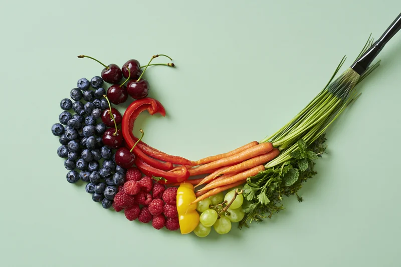 Rainbow arc of fresh fruits and vegetables arranged in color order on a light green background with a paintbrush forming the tail
