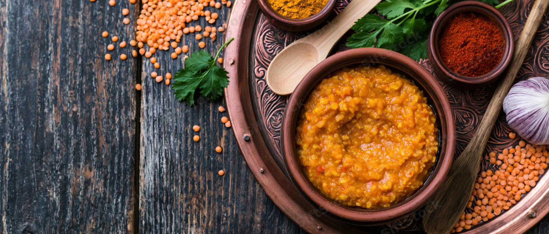 Red lentil dal in a clay bowl on an engraved copper tray with spices and herbs on a dark wooden table