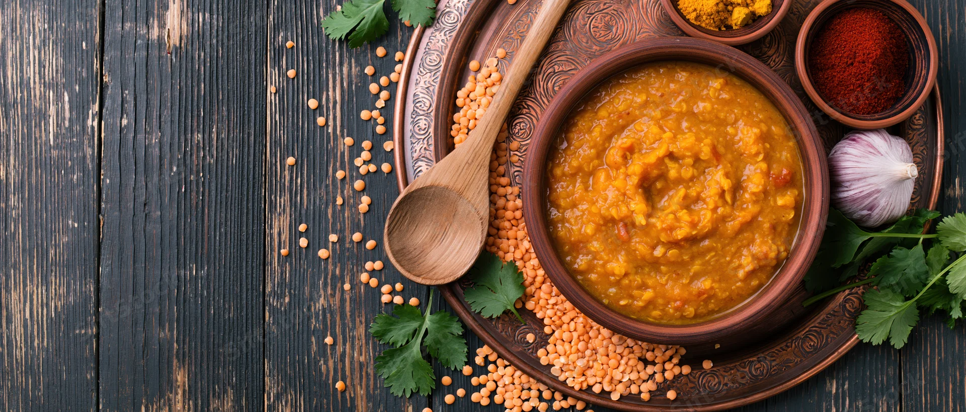 Red lentil dal in a clay bowl on an engraved copper tray with spices and herbs on dark wood