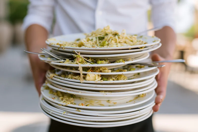Server holding tall stack of used white dinner plates with food remnants and forks outdoors in daylight