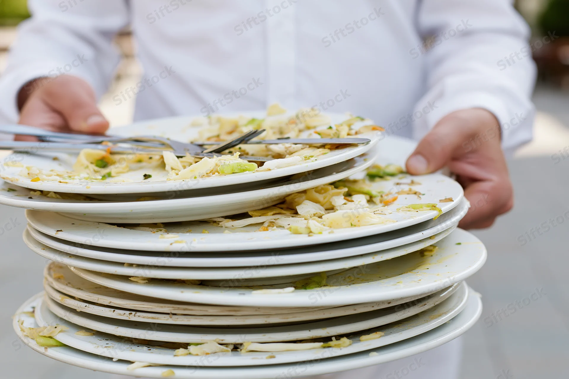 Server holding a tall stack of used white dinner plates with crumbs and utensils