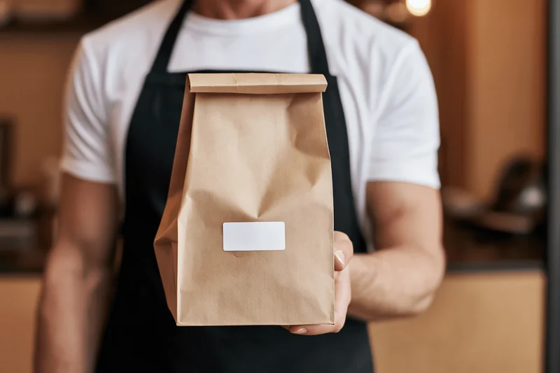 Man in black apron holding brown paper bag with blank label in restaurant interior