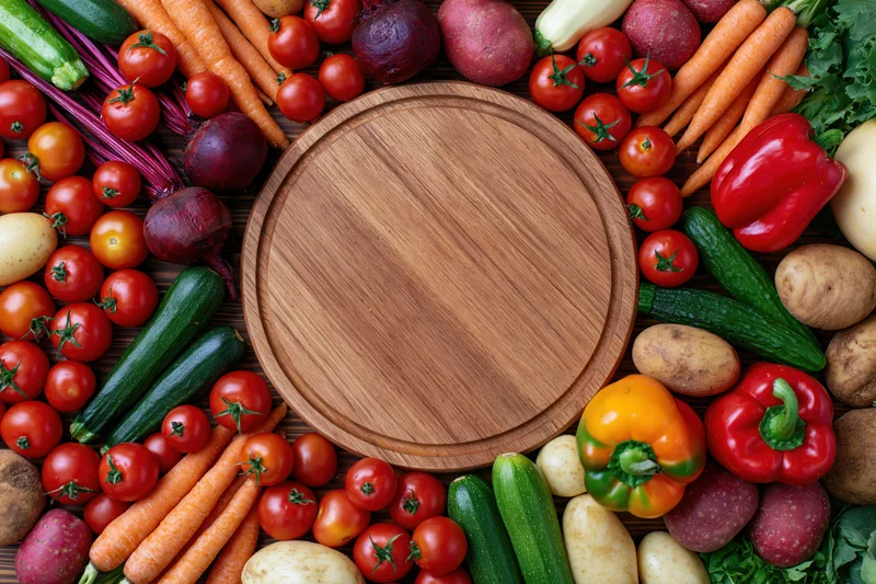 Round wooden cutting board surrounded by fresh vegetables on dark wood table