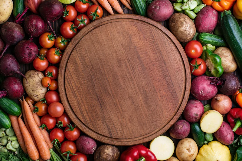Round wooden cutting board surrounded by fresh vegetables on dark wood surface