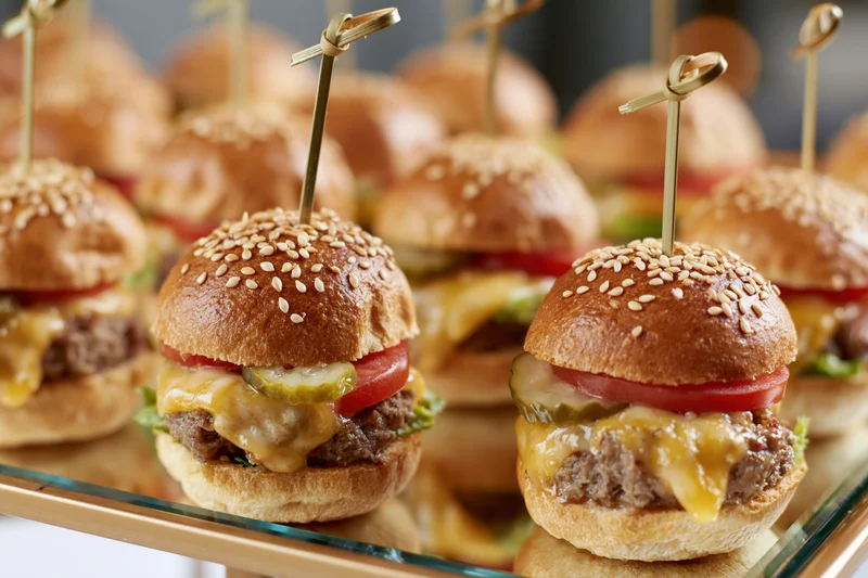 Mini beef sliders with cheese, tomato, and pickle on sesame buns arranged on a mirrored glass tray