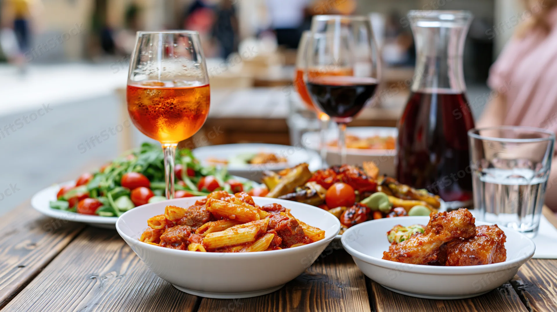 Pasta dish with meatballs and tomato sauce served on a rustic wooden outdoor table alongside salad, chicken wings, wine, and water