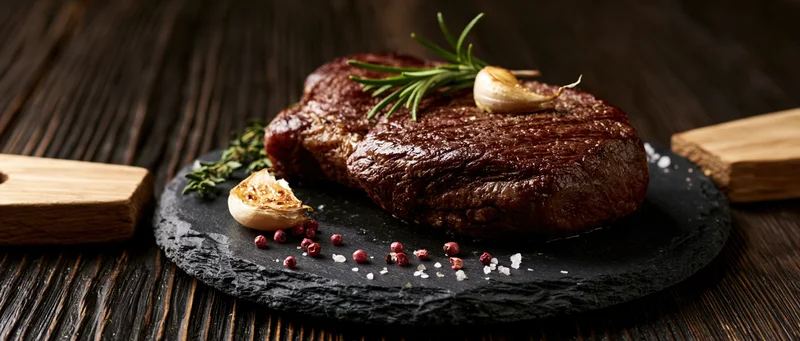 Seared beef steak on dark slate board with rosemary and garlic, surrounded by pepper and salt on wooden table