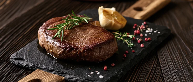 Seared beef steak on dark slate board with herbs and garlic, wooden table background