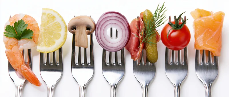 Seven forks each holding a different food item arranged in a row on a white surface