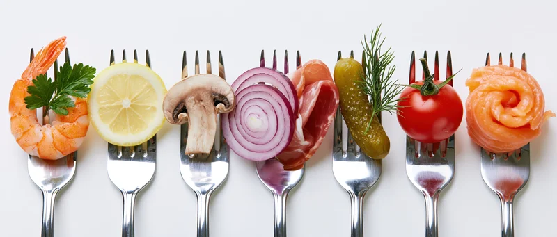 Seven forks each holding a different food item arranged in a row on a white surface