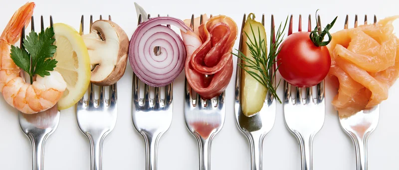 Seven forks each holding a different food item arranged in a row on a white surface