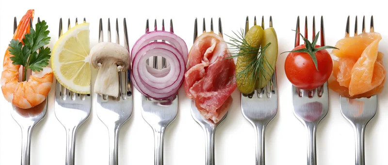 Seven forks each holding a different appetizer ingredient arranged in a row on a white background