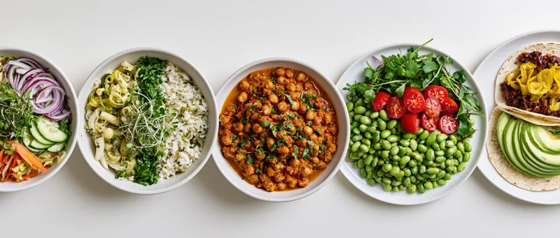 Five plant-based meals in white bowls and plates arranged in a row on a white surface