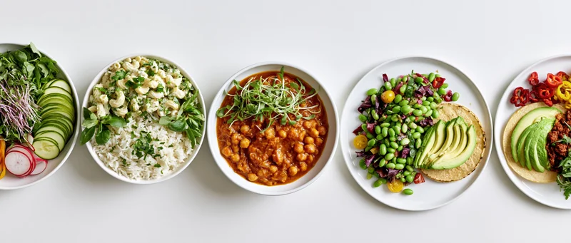 Five plant-based meals served in white bowls and plates arranged in a horizontal row on a white surface