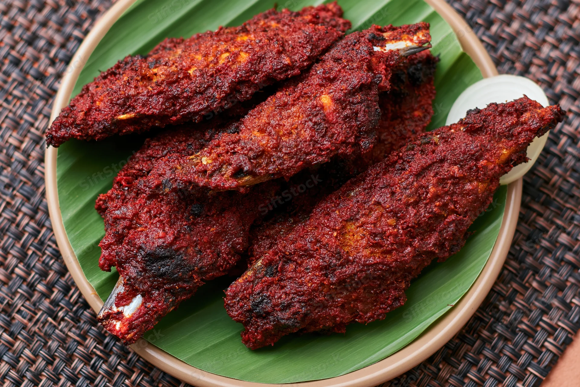 Deep-fried spiced fish pieces coated in dark red curry sauce served on banana leaf in bowl on woven mat