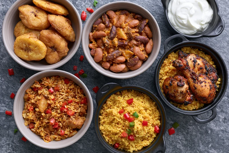 Six bowls of west african food arranged on a dark slate surface with rice, beans, plantain, chicken, couscous and yogurt