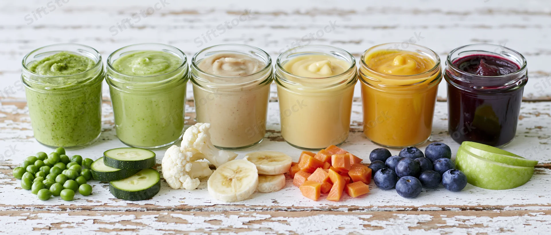 Six glass jars of colorful baby food puree arranged in a row on a rustic wooden surface with fresh fruits and vegetables in front