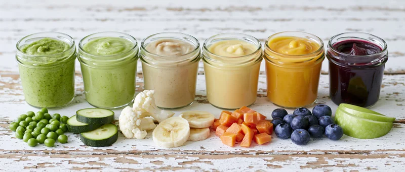 Six glass jars of colorful baby food puree arranged in a row on a rustic wooden surface with fresh fruits and vegetables in front