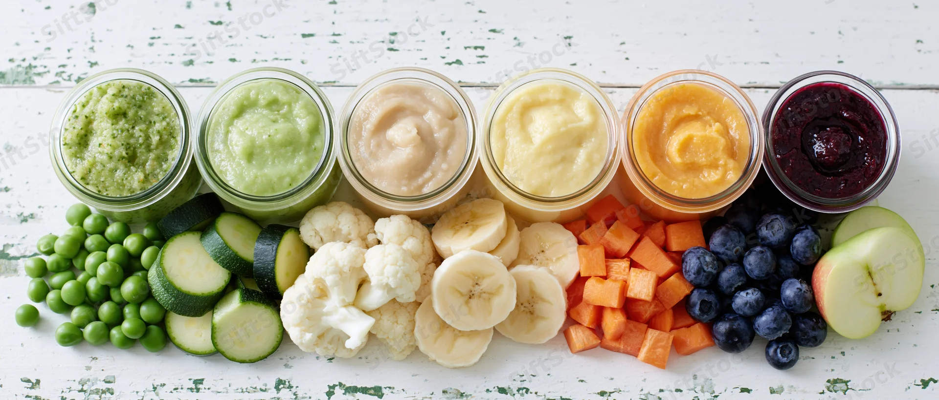 Six glass jars of colorful baby food puree arranged in a row with fresh vegetables and fruits on a white wooden surface