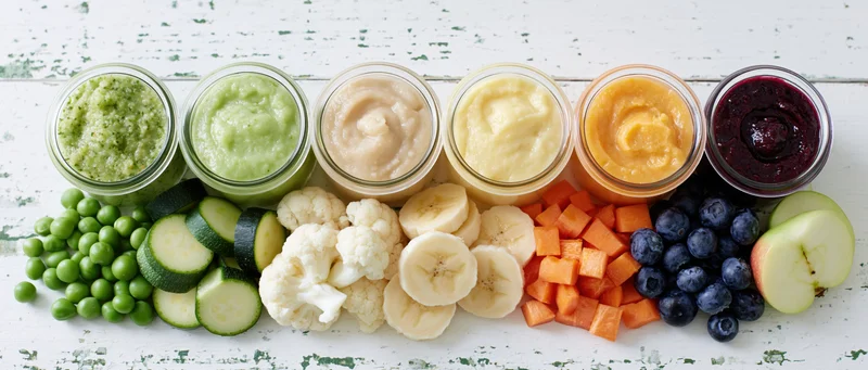 Six glass jars of colorful baby food puree arranged in a row with fresh vegetables and fruits on a white wooden surface