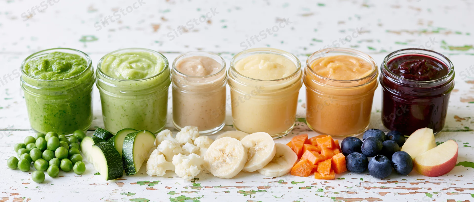 Six glass jars of colorful baby food puree arranged in a row with fresh fruits and vegetables on a white rustic wooden surface