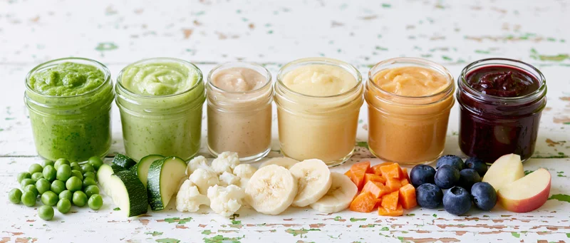 Six glass jars of colorful baby food puree arranged in a row with fresh fruits and vegetables on a white rustic wooden surface