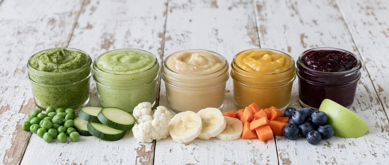 Six glass jars of colorful baby food puree arranged in a row on a white wooden table with fresh vegetables and fruits in front
