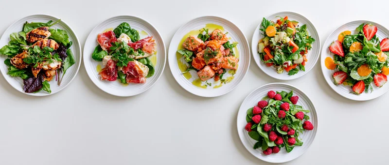 Six white ceramic plates with assorted salads and garnishes arranged in a row on a white surface