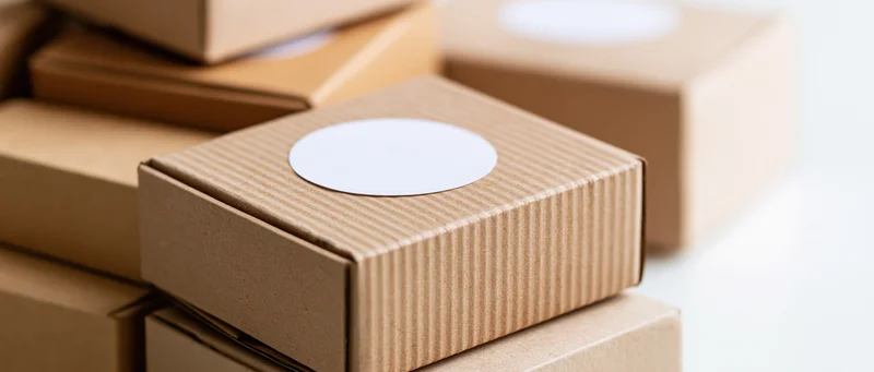 Small kraft cardboard box with white circular sticker stacked among others on plain surface