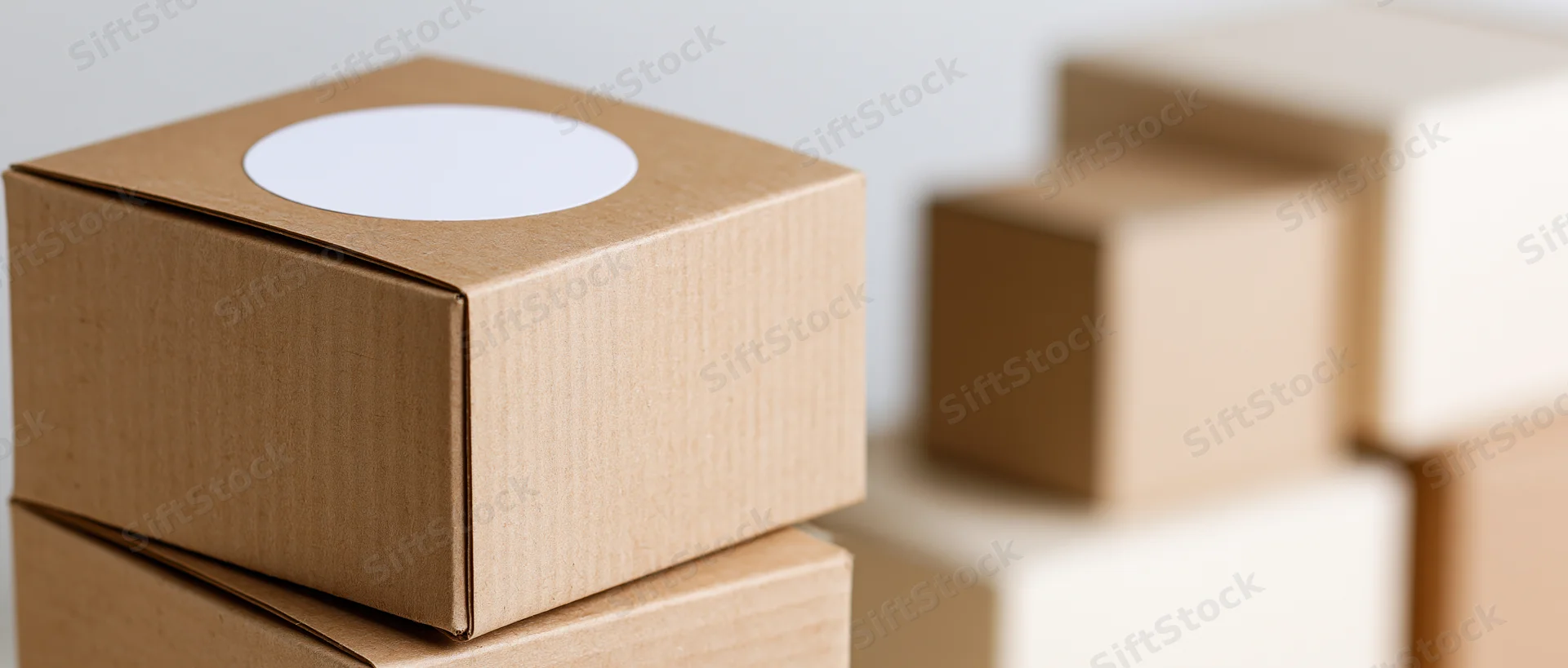 Small kraft cardboard box with white circular sticker stacked on another box, with blurred boxes in background