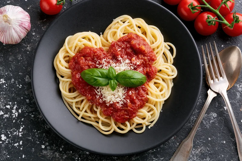 Spaghetti with tomato sauce arranged in heart shape on black plate with basil and cheese, surrounded by garlic and tomatoes, cutlery on dark surface
