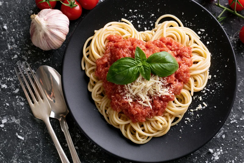 Spaghetti with tomato sauce arranged in heart shape on black plate with basil and cheese, surrounded by garlic and tomatoes, cutlery on dark stone surface