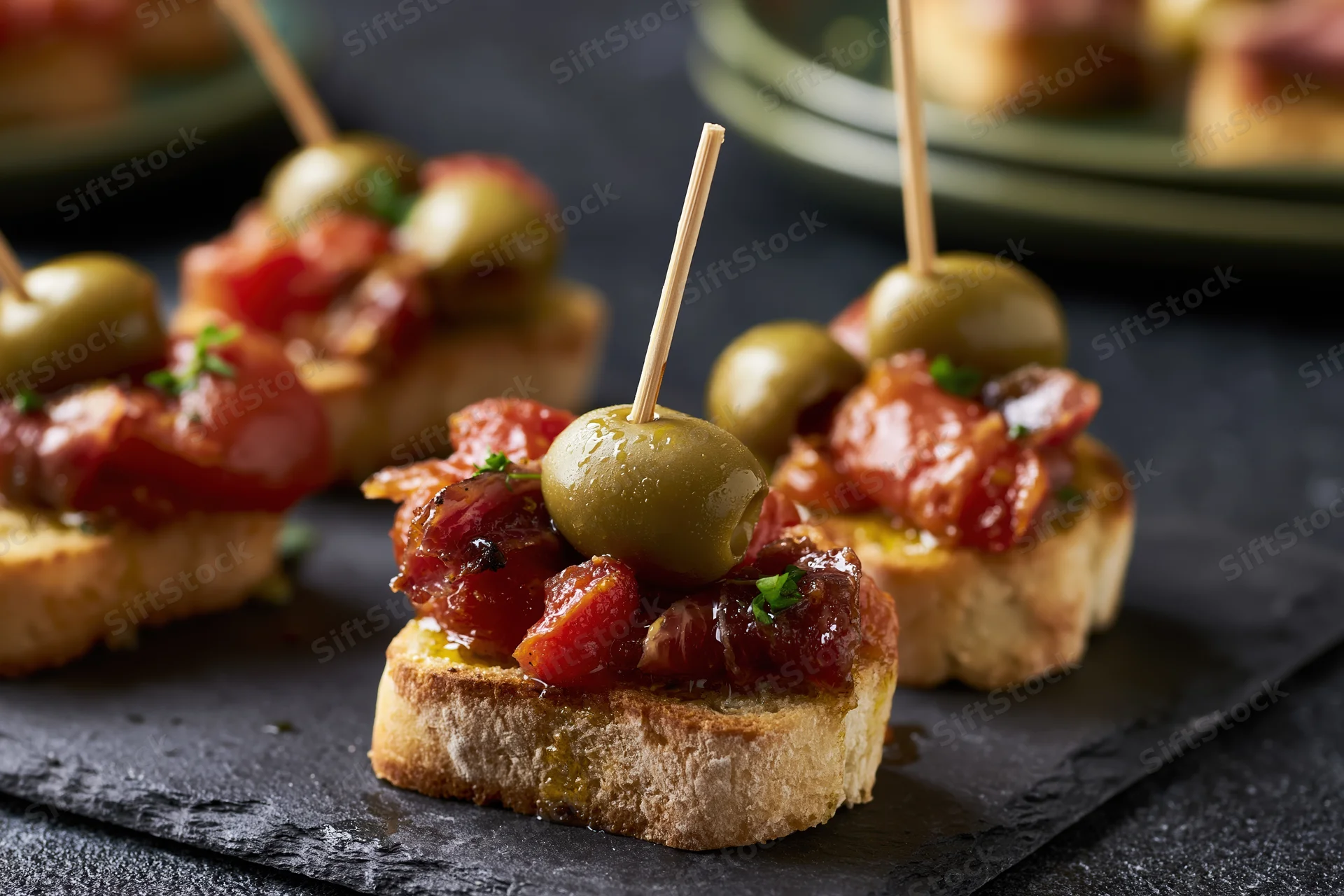 Spanish pintxos on toasted baguette with olives and jamon on dark slate plate