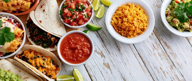 Spread of mexican dishes including tacos, rice, salsa, guacamole, and lime wedges arranged along the left edge of a white wooden table