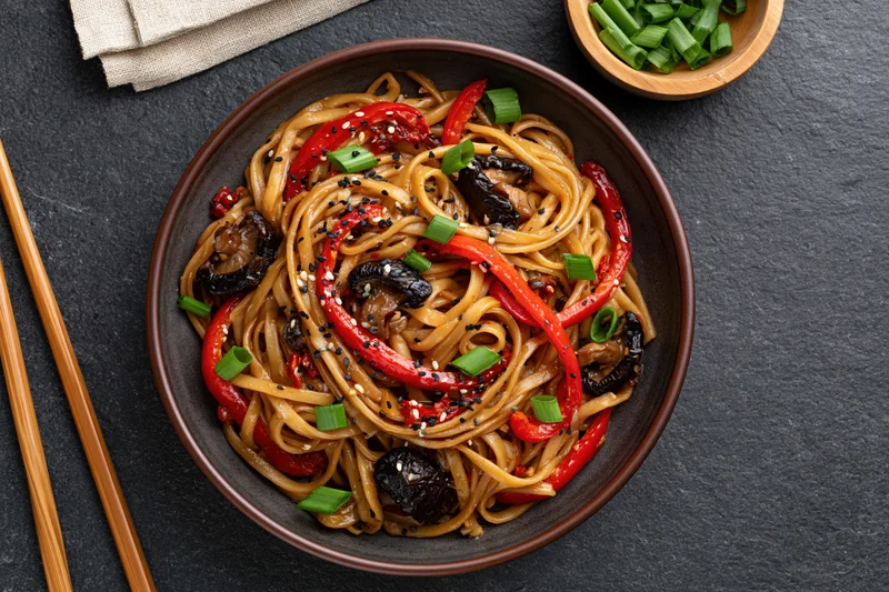 Stir-fried noodles with red bell peppers and shiitake mushrooms in a bowl, garnished with green onions and sesame seeds, served with chopsticks on a dark slate surface