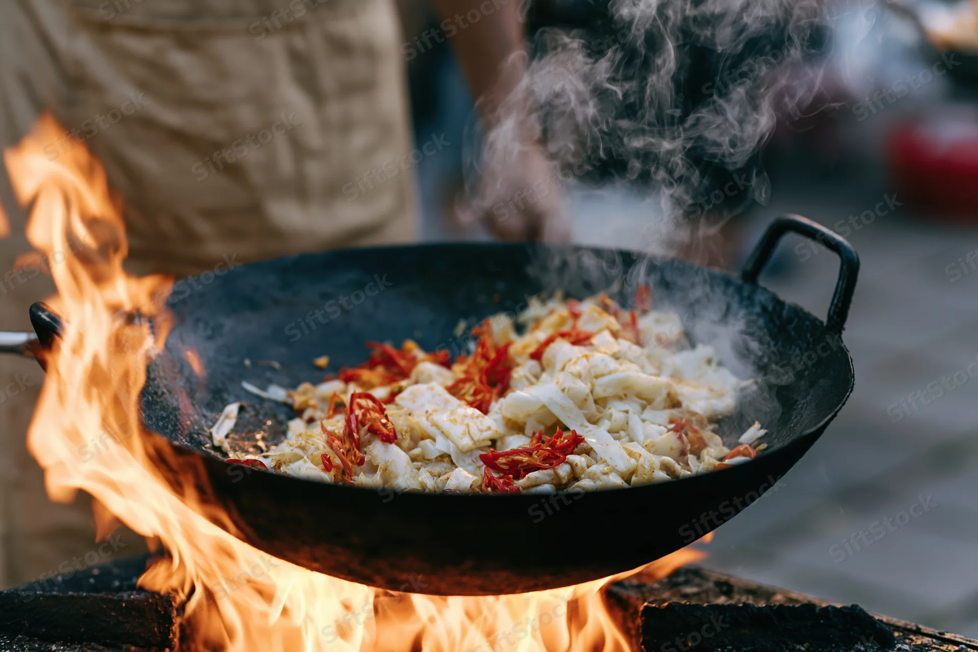 Chef stir-frying vegetables and chili in a wok over open flames outdoors