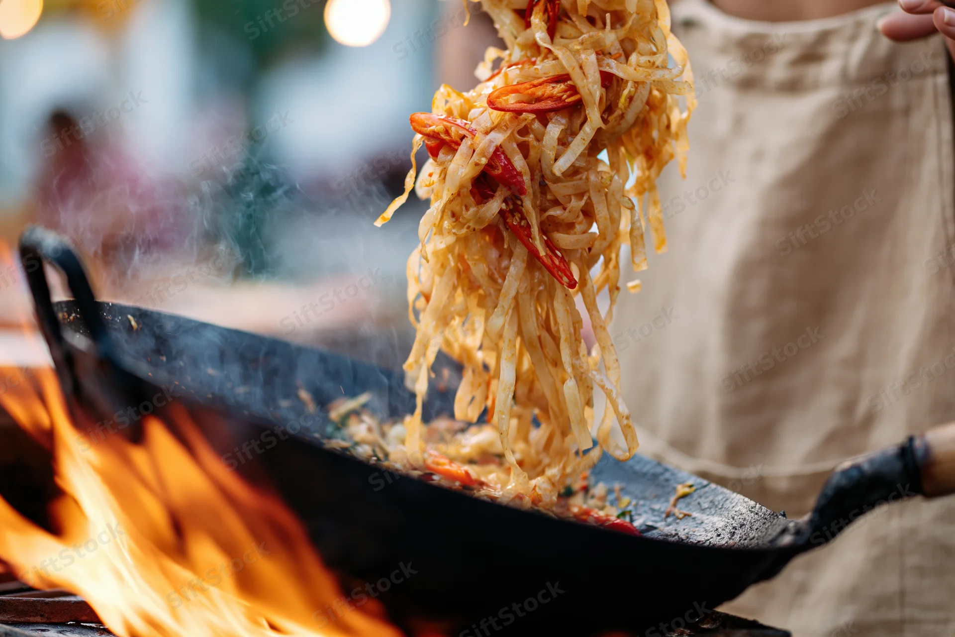 Cook tossing rice noodles with red chili peppers in a wok over open flame at street market