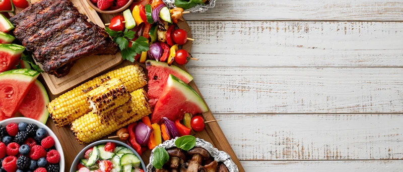 Summer barbecue spread with grilled ribs, corn, watermelon and vegetables arranged on wooden board along left side of white table